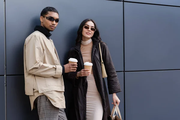 Hombre afroamericano y mujer joven en gafas de sol con estilo sosteniendo vasos de papel y bolsa de compras - foto de stock