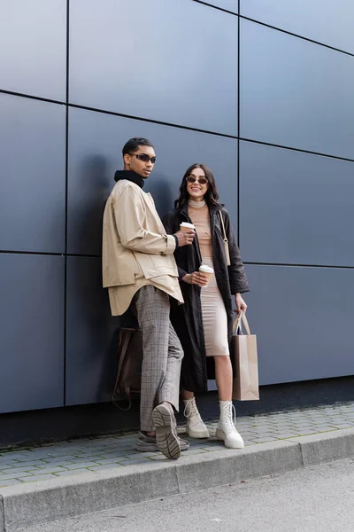 Élégant homme afro-américain et heureuse jeune femme dans des lunettes de soleil tenant des tasses en papier et un sac à provisions — Photo de stock