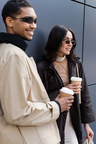 Hombre afroamericano feliz y mujer joven en gafas de sol con estilo sosteniendo vasos de papel afuera - foto de stock