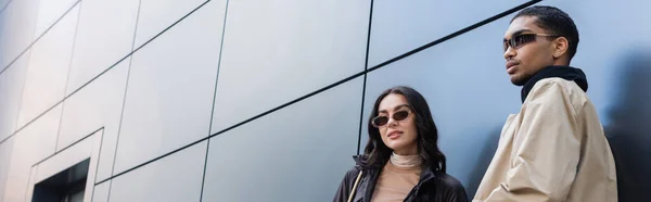 Elegante pareja multiétnica y joven en gafas de sol cerca del edificio, pancarta - foto de stock