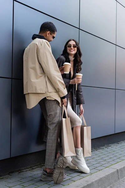 Homem americano africano alegre e jovem mulher em óculos de sol elegantes segurando copos de papel e sacos de compras — Fotografia de Stock