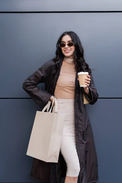 Mujer joven feliz en traje elegante y gafas de sol sosteniendo taza de papel y bolsa de compras cerca del edificio - foto de stock