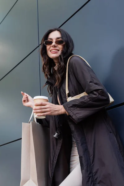 Glückliche brünette Frau in stylischem Outfit und Sonnenbrille mit Pappbecher und Einkaufstasche in der Nähe des Gebäudes — Stockfoto