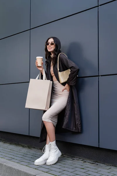 Femme brune en tenue élégante et lunettes de soleil tenant tasse en papier et sac à provisions tout en souriant près du bâtiment — Photo de stock