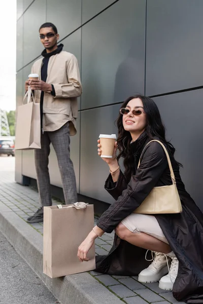 Giovane donna alla moda con borsetta e tazza di carta vicino alla borsa della spesa e l'uomo afroamericano offuscato — Foto stock
