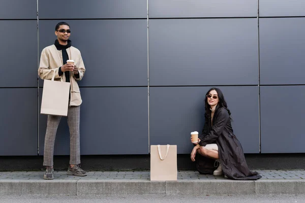 Молодая стильная женщина с бумажной чашкой сидит рядом с сумкой и молодой африканский американец — стоковое фото