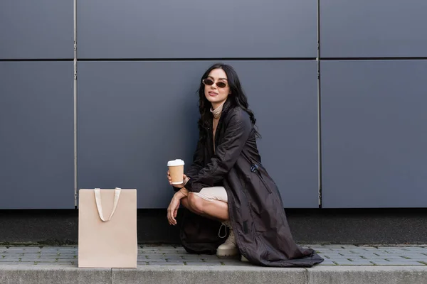 Mujer joven y elegante con taza de papel sentado cerca de la bolsa de papel y la construcción de centro comercial - foto de stock