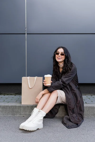 Jovem mulher feliz com copo de papel sentado perto de saco de papel e construção de shopping center — Fotografia de Stock