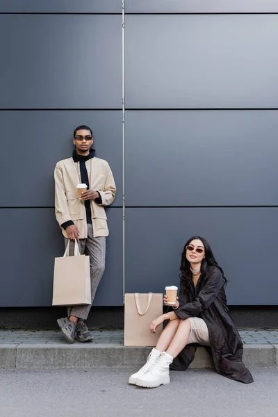 Jovem mulher com copo de papel sentado perto de saco de compras e elegante homem afro-americano — Fotografia de Stock