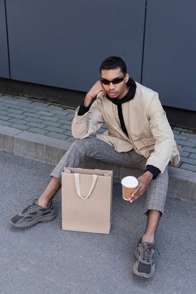 Vista ad alto angolo dell'uomo afroamericano in occhiali da sole e vestito autunnale seduto con tazza di carta vicino alla borsa della spesa — Foto stock