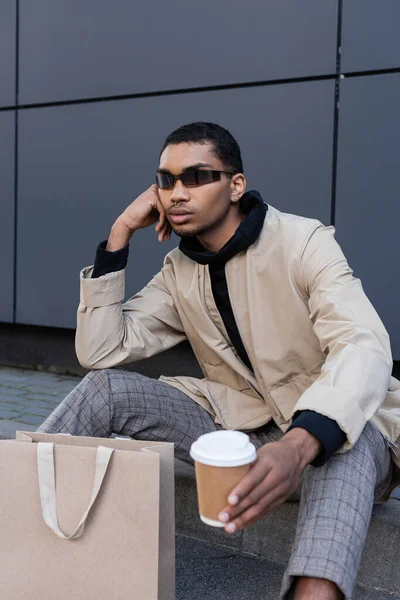 Hombre afroamericano en gafas de sol y traje otoñal sentado con café para ir cerca de la bolsa de compras - foto de stock