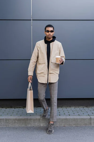Comprimento total do homem americano africano elegante em óculos de sol e outfit outfit segurando copo de papel e saco de compras — Fotografia de Stock