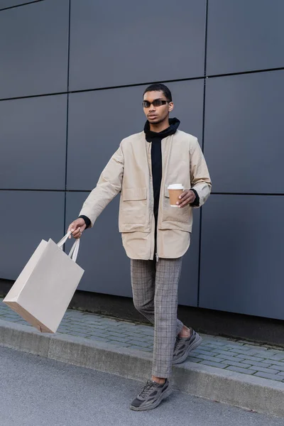 Longitud completa de hombre afroamericano con estilo en gafas de sol y traje otoñal sosteniendo taza de papel y caminando con bolsa de compras - foto de stock