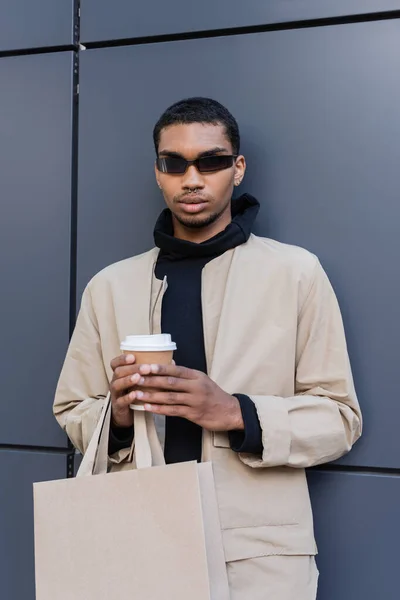 Stylish african american man in sunglasses and autumnal outfit holding paper cup and shopping bag — Stock Photo