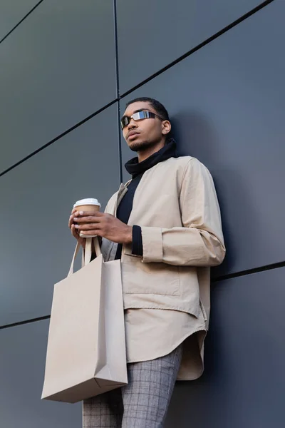 Baixo ângulo vista de homem americano africano elegante em óculos de sol e outfit outfit segurando copo de papel e saco de compras — Fotografia de Stock