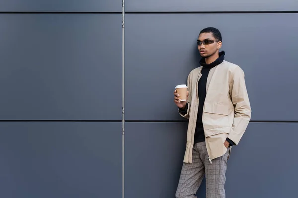 Hombre afroamericano con estilo en gafas de sol y traje otoñal sosteniendo taza de papel y de pie con la mano en el bolsillo - foto de stock