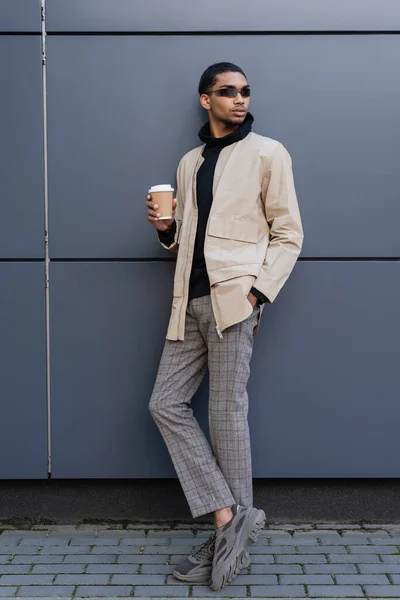 Hombre afroamericano con estilo en elegante traje otoñal sosteniendo taza de papel y de pie con la mano en el bolsillo — Stock Photo