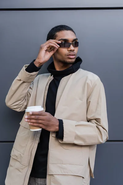 Homem americano africano elegante na roupa outnal segurando copo de papel e ajustando óculos de sol — Fotografia de Stock