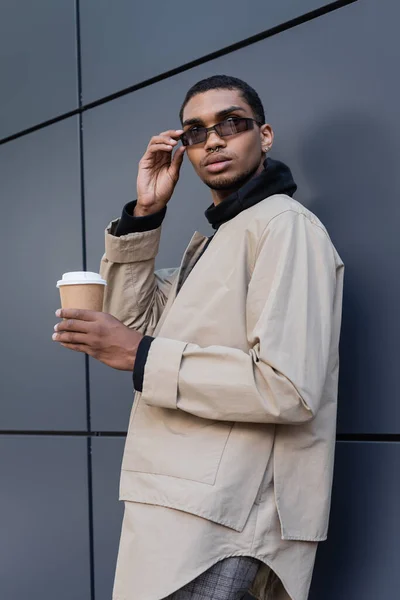 Stylish african american man in autumnal outfit holding paper cup and adjusting sunglasses near building — Stock Photo