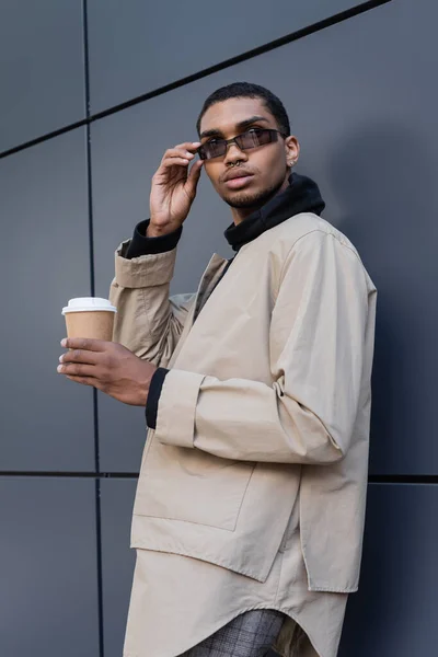 Hombre afroamericano con estilo en traje otoñal sosteniendo taza de papel y ajustando gafas de sol cerca del edificio — Stock Photo