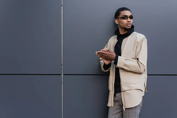 Молодий афроамериканець в автумному одязі і стилізовані сонячні окуляри стоять поруч з будинком. — стокове фото
