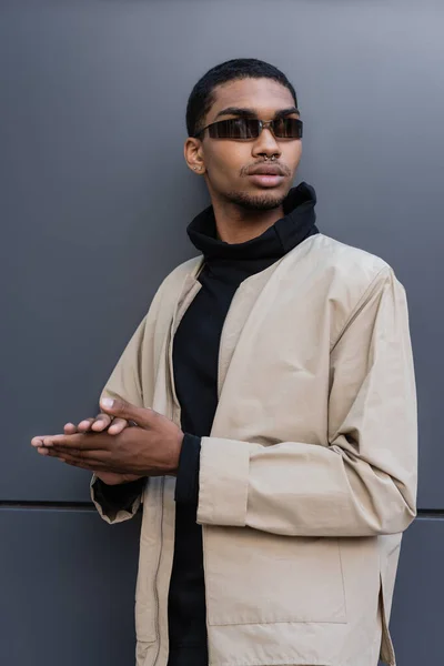 Confiant jeune homme afro-américain en tenue automnale et des lunettes de soleil élégantes debout près du bâtiment — Photo de stock