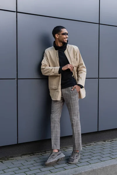 Homem americano africano feliz na roupa outnal e óculos de sol elegantes que estão com a mão no bolso perto do edifício — Fotografia de Stock