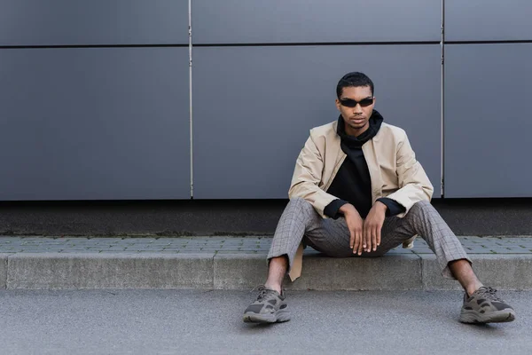 In voller Länge junger afrikanisch-amerikanischer Mann in herbstlichem Outfit und stylischer Sonnenbrille sitzt in der Nähe des Gebäudes — Stockfoto