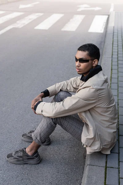 Comprimento total de jovem afro-americano em roupa outnal e óculos de sol elegantes sentados na rua urbana — Fotografia de Stock