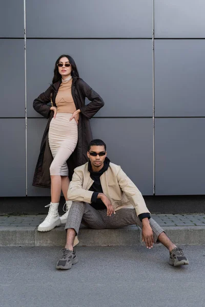 Longitud completa de elegante pareja multiétnica en trajes de otoño de moda y gafas de sol posando cerca de la construcción - foto de stock