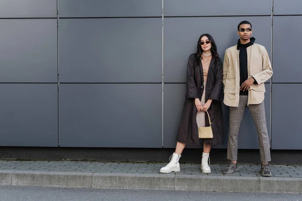 Longitud completa de la joven pareja multiétnica en trajes otoñales de moda y gafas de sol posando cerca de la construcción - foto de stock