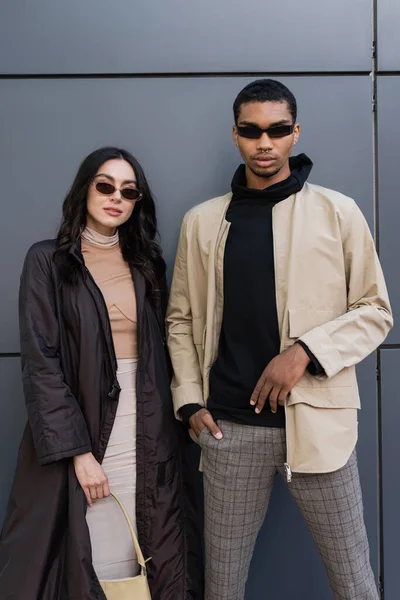 Giovane coppia multietnica in abiti autunnali alla moda e occhiali da sole in posa vicino edificio — Foto stock