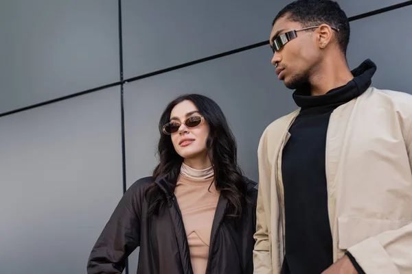 Vista de ángulo bajo de la elegante pareja multiétnica en trajes otoñales de moda y gafas de sol de pie cerca del edificio - foto de stock