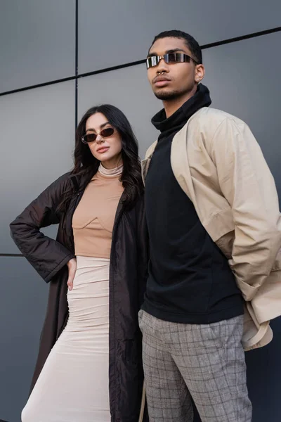 Elegante pareja multiétnica en trajes otoñales y gafas de sol de moda de pie cerca del edificio - foto de stock