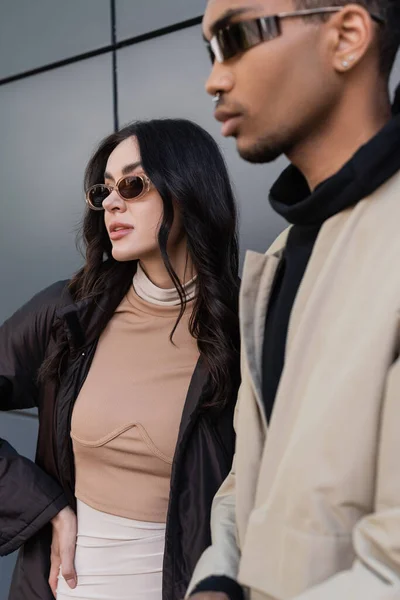 Mujer joven con estilo en traje otoñal de moda y gafas de sol de pie cerca del hombre afroamericano en primer plano borroso - foto de stock