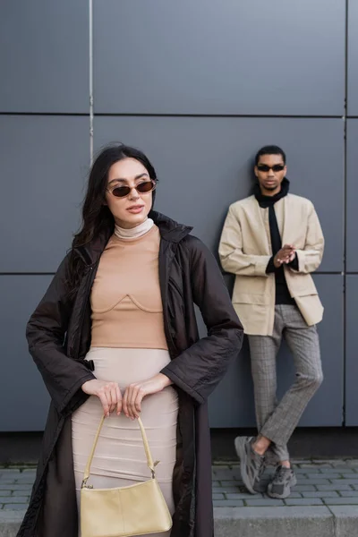 Mujer joven con estilo en traje otoñal y gafas de sol de moda de pie cerca del hombre afroamericano sobre fondo borroso - foto de stock