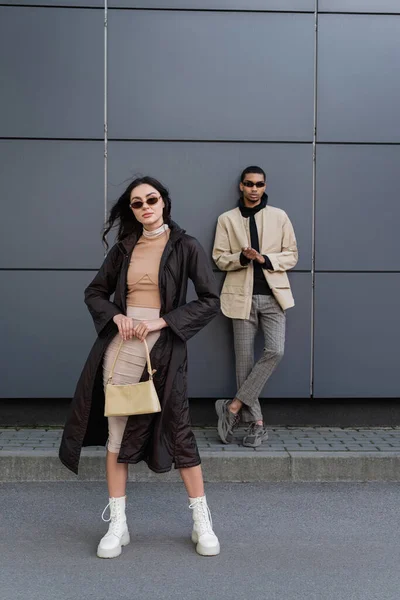 Comprimento total da jovem mulher elegante em casaco outonal e óculos de sol de pé com bolsa perto de homem americano africano — Fotografia de Stock