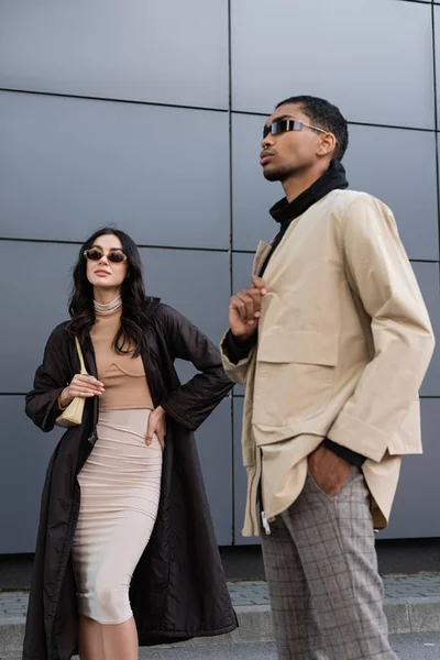 Pareja multiétnica con elegantes trajes de otoño y gafas de sol posando cerca del edificio - foto de stock