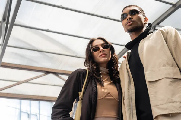 Vista de ángulo bajo de la mujer con estilo con bolso de mano en gafas de sol de moda mirando a la cámara cerca de hombre afroamericano - foto de stock