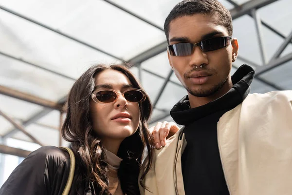 Retrato de casal interracial elegante em óculos de sol da moda olhando para a câmera — Fotografia de Stock