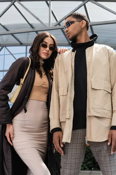 Low angle view of stylish interracial couple in sunglasses and stylish outfits looking at camera — Stock Photo