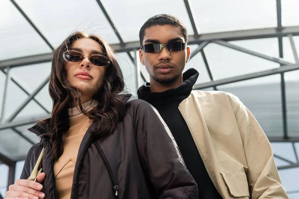 Vue à angle bas de couple élégant multiethnique dans des lunettes de soleil regardant la caméra — Photo de stock