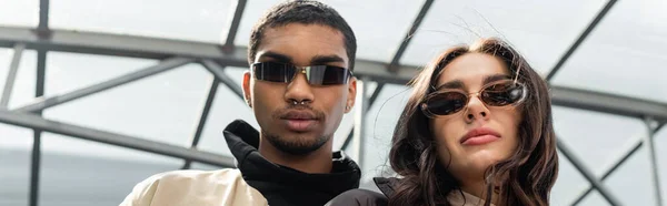 Low angle view of stylish interracial couple in sunglasses looking at camera, banner — Stock Photo