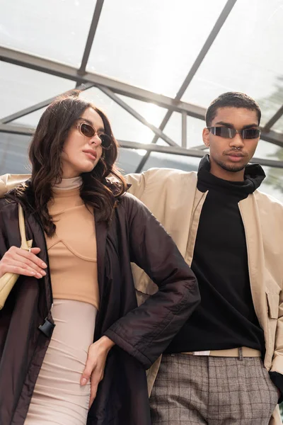 Vista de ángulo bajo de elegante pareja interracial en gafas de sol posando bajo el techo - foto de stock