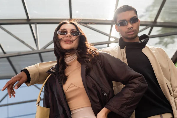 Baixo ângulo de visão de casal interracial elegante em óculos de sol olhando para a câmera — Fotografia de Stock