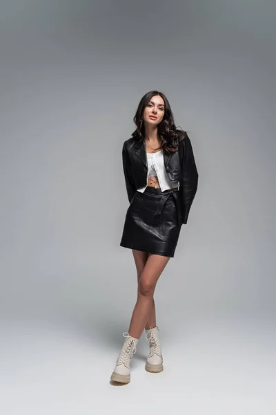 Full length of young woman in stylish black leather jacket and boots posing on grey — Stock Photo