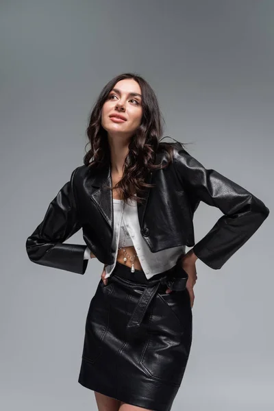 Dreamy young woman in stylish black leather jacket and skirt posing with hands on hips isolated on grey — Stock Photo