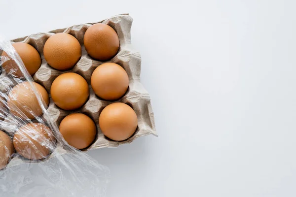 Vista superior de ovos castanhos em bandeja de cartão com celofane sobre fundo branco — Fotografia de Stock