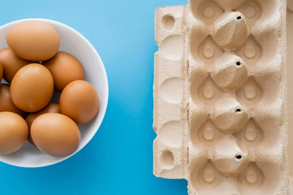 Top view of eggs in bowl near carton container on blue background — Stock Photo