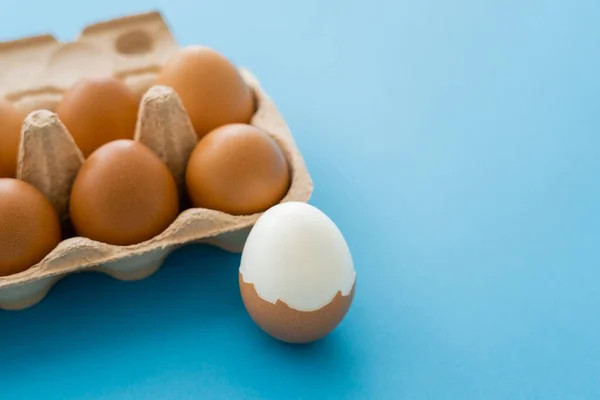 Вареные и сырые яйца в раковинах рядом с упаковкой картона на синем фоне — стоковое фото